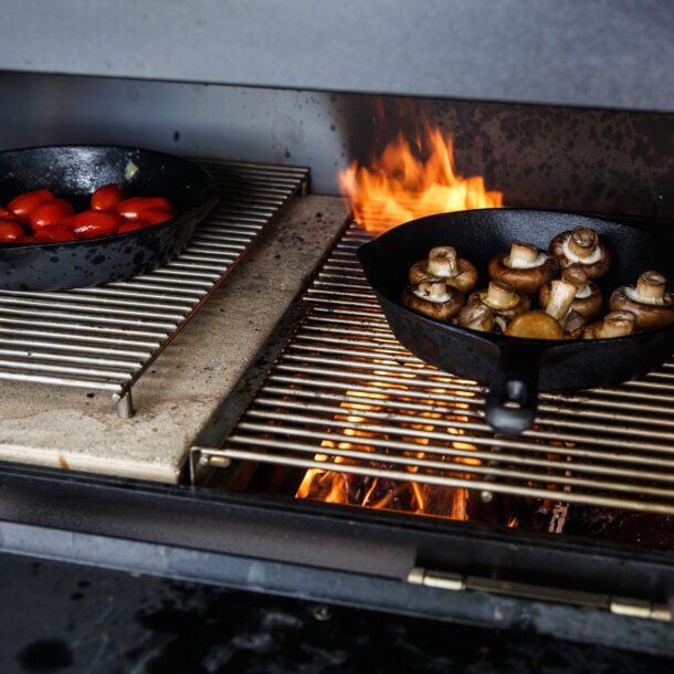 Zwei gusseiserne Pfannen mit Champignons und Kirschtomaten auf doppeltem Grillrost.“