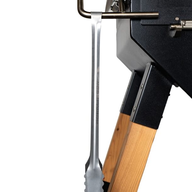  „MERKLINGER Grillzange seitlich am Ofen eingehängt, Gerätefüße aus Holz sichtbar.“