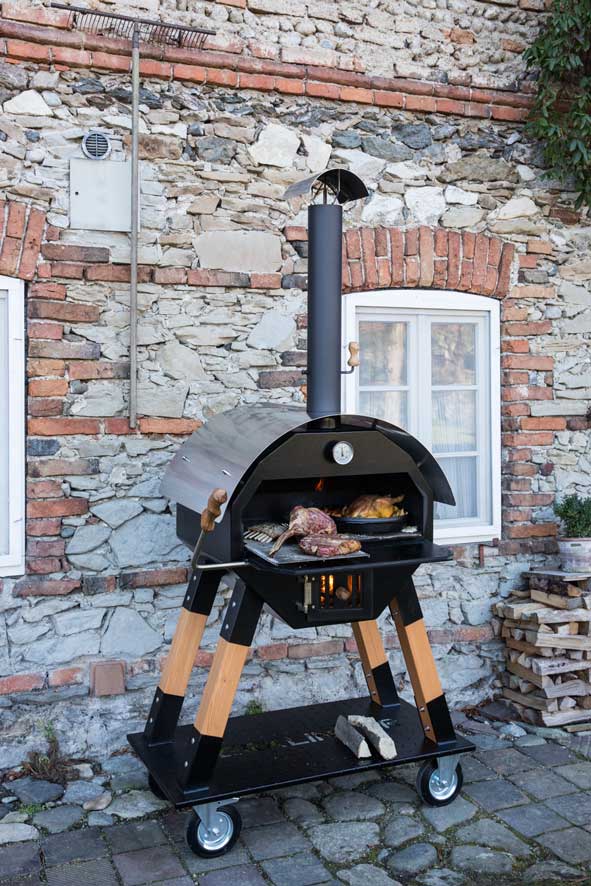 Mobiler MERKLINGER Rundofen auf Rollen vor einer rustikalen Steinmauer Merklinger Holzbackofen Outdoor mit Holzfeuer – Grillen und Backen vor rustikaler Natursteinwand, gefüllt mit Fleisch und Beilagen