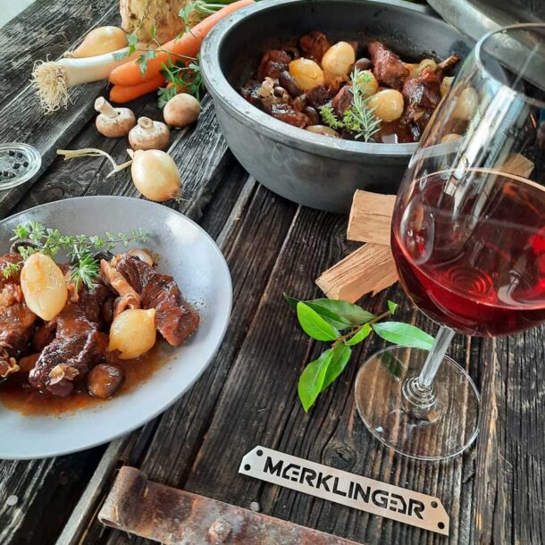 Schmorgericht & Wein: Rustikaler Tisch mit Teller und Topf voller geschmorter Fleischstücke und Perlzwiebeln; Rotweinglas daneben, Gemüse und Kräuter als Dekoration; Merklinger-Logo im Vordergrund