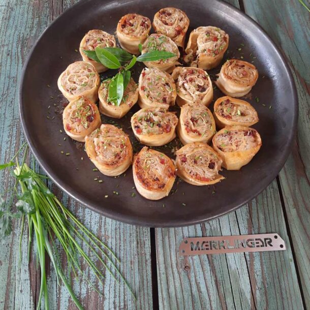 Rustikale Draufsicht: Eine runde dunkle Platte mit herzhaften Teigschnecken, mit Kräutern bestreut; daneben frische Kräuterzweige und ein „MERKLINGER“-Metallschild Merklinger Zubehör Denkkeramik Keltenschale 05