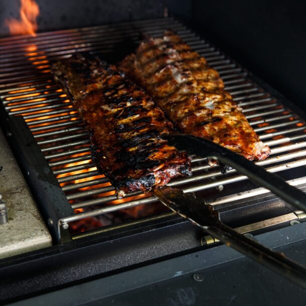 Würzige Spare Ribs auf Edelstahl-Grillrost mit Flammen im Hintergrund.“