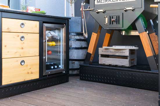 Merklinger Outdoor-Küchenmodul mit Glasfront-Kühlschrank und drei Schubladen neben MERKLINGER Ofen auf Terrasse.