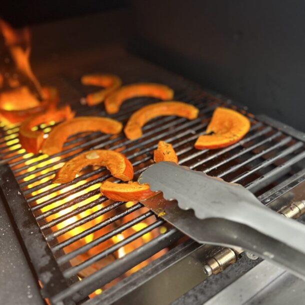 „MERKLINGER Grillzange greift gegrillte Kürbisspalten auf dem Rost, Flammen im Hintergrund.“