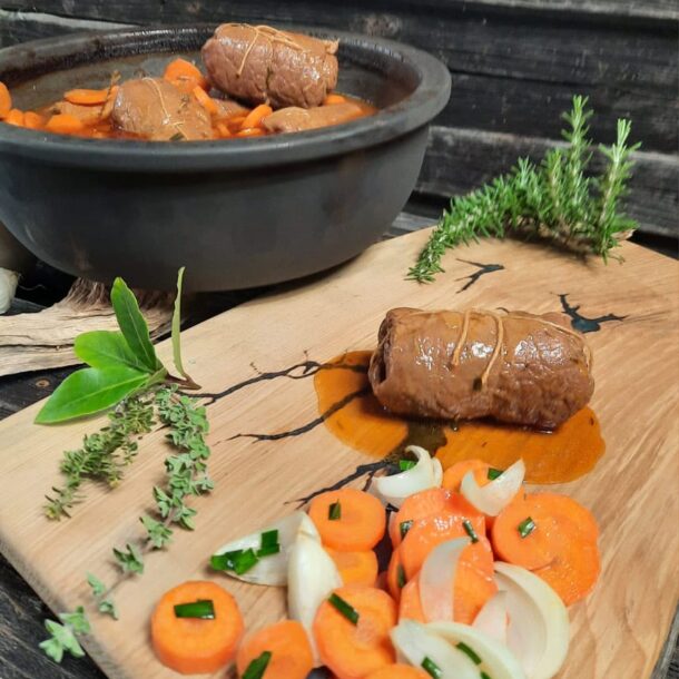 Rouladen im Topf: Keramiktopf mit Rouladen in Bratensauce; vorne eine Roulade auf Holzbrett mit Karotten- und Zwiebelstücken sowie Kräuterzweigen