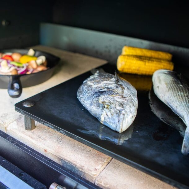 Ganze Fische auf schwarzer Plancha im Merklinger Ofen, daneben Maiskolben und eine Pfanne mit Gemüse