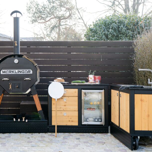Modulare MERKLINGER Outdoor-Küche mit Holzbackofen und Granit-Arbeitsplatte – Familie bereitet Zutaten vor.“