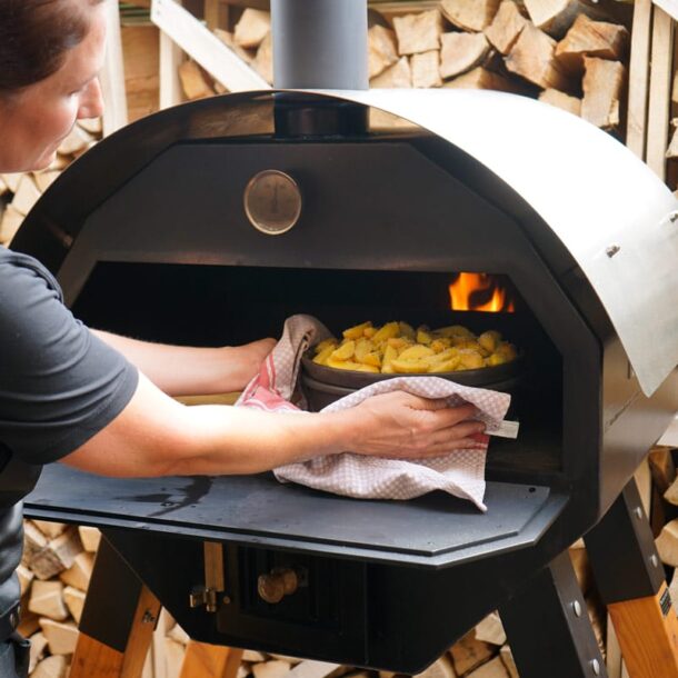 Backen im Holzofen: Eine Person schiebt eine Auflaufform mit Kartoffeln in den befeuerten Merklinger-Holzbackofen; Holzstapel im Hintergrund.