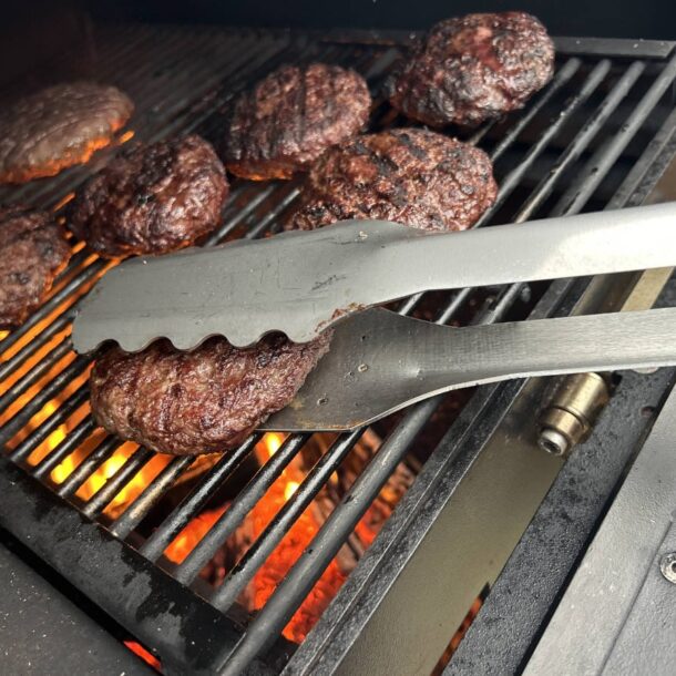  „MERKLINGER Grillzange wendet ein Burgerpatty auf dem Grillrost über offener Flamme.“