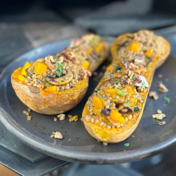 Zwei halbierte und gefüllte Butternut-Kürbisse auf einer dunklen Platte – die Füllung aus Getreidekörnern und Gemüse, teils mit Kräutern bestreut. DerMerklinger_Holzbackofen_Zubehoer_Denkkeramik_Keltenschale_04-qenup6dy4hsrue7cudwv0isjld7sfvr3tgf9ue6hjo