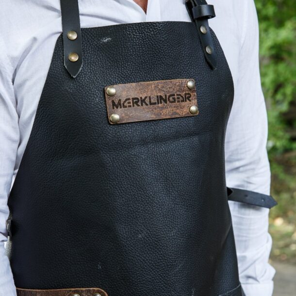  „MERKLINGER Lederschürze getragen – Brustbereich mit Logo, verstellbare Lederriemen, weiße Bluse im Hintergrund.“