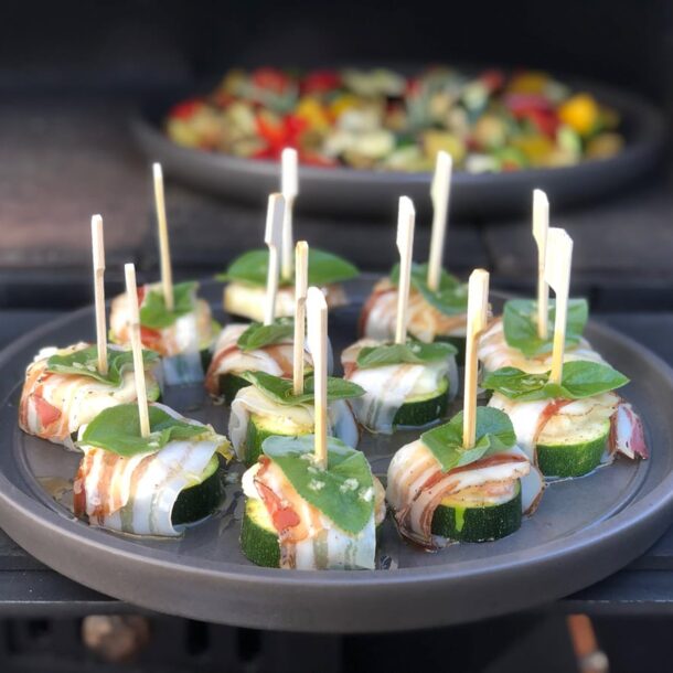 Fingerfood im Ofen: Zucchini-Speck-Röllchen auf Keramikplatte, mit Holzspießen fixiert; dahinter eine zweite Platte mit buntem Grillgemüse.
