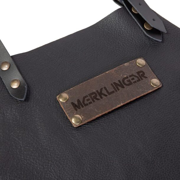 „Geprägtes MERKLINGER Logo auf brauner Lederplakette mit Messingnieten – Detail der schwarzen Lederschürze.“