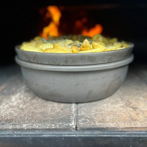 Doppelschale im Ofen: Zwei ineinander gestellte, runde Keramikformen (Merklinger-Zubehör) mit Kartoffelgratin im heißen Holzbackofen, Flammen im Hintergrund.