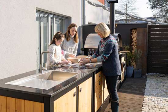 Drei Generationen arbeiten am langen Spülmodul; MERKLINGER Ofen im Hintergrund auf Terrasse.