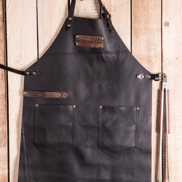 MERKLINGER Lederschürze flach auf Holzuntergrund – zwei Taschen, Werkzeugleiste, Grillzange seitlich eingehängt.“