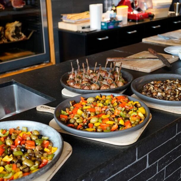 Buffet-/Showküchenszene: Mehrere runde Ofenplatten auf Holzbrettern mit Ofengemüse, Pilzen und Fleischspießen, arrangiert auf einer schwarzen Arbeitsfläche. Merklinger Zubehör Denkkeramik Keltenschale 2 (4 Stück) mit Essen