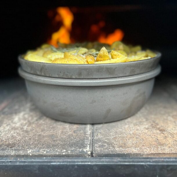 Doppelschale im Ofen: Zwei ineinander gestellte, runde Keramikformen (Merklinger-Zubehör) mit Kartoffelgratin im heißen Holzbackofen, Flammen im Hintergrund.