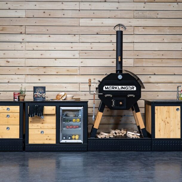  „Komplette MERKLINGER Outdoor-Küche im Showroom mit Holz-Stahl-Modulen, Kühlschrank mit Glastür und Holzbackofen.“