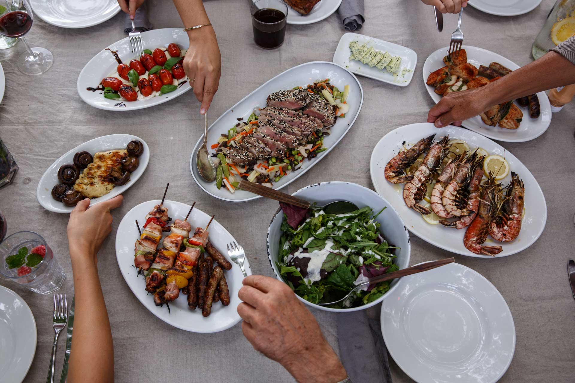 Blick von oben auf einen Tisch mit mehreren Tellern: gegrillte Garnelen, Spieße, Salat, Tomaten und Pilze.