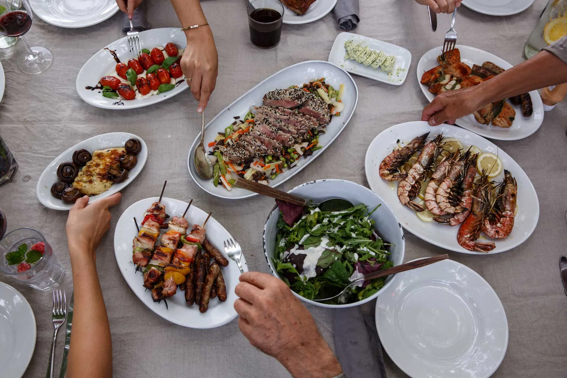 Blick von oben auf einen Tisch mit mehreren Tellern: gegrillte Garnelen, Spieße, Salat, Tomaten und Pilze.