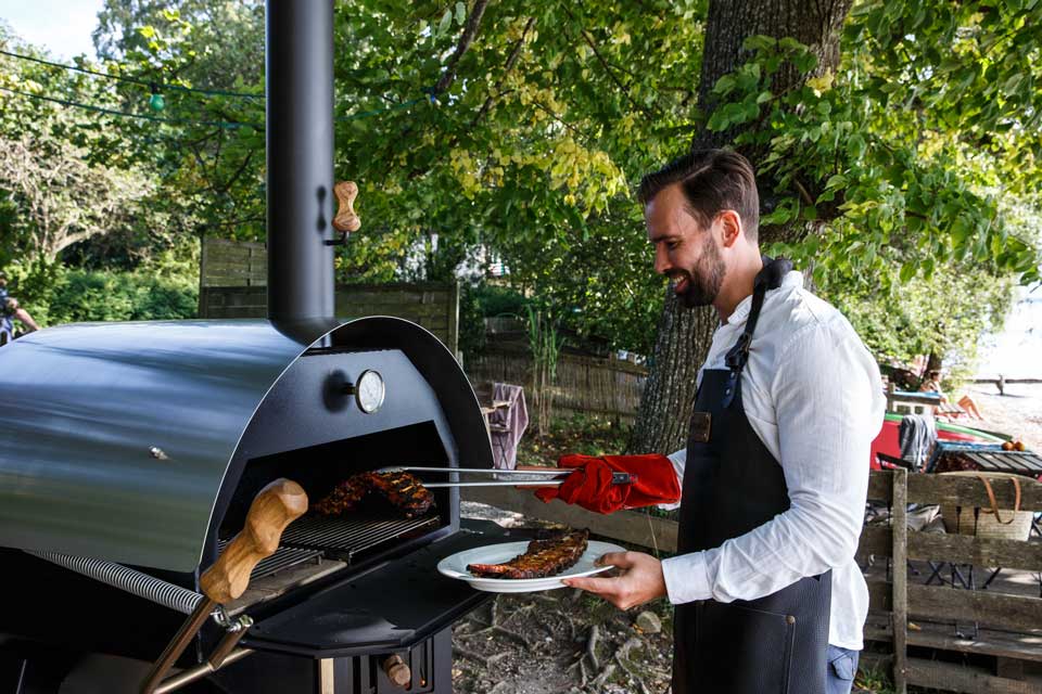 Mann in Schürze entnimmt gegrillte Rippchen mit Zange aus MERKLINGER Rundofen im Garten