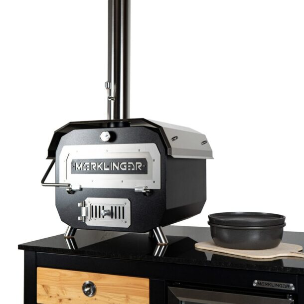 Merklinger-Ofen auf Arbeitsplatte: Kompakter, schwarzer Holzbackofen von Merklinger mit Edelstahlfront und Ofenrohr; daneben gusseiserne Form auf Holzbrett. Merklinger Bobby Outdoor Keramik Modell