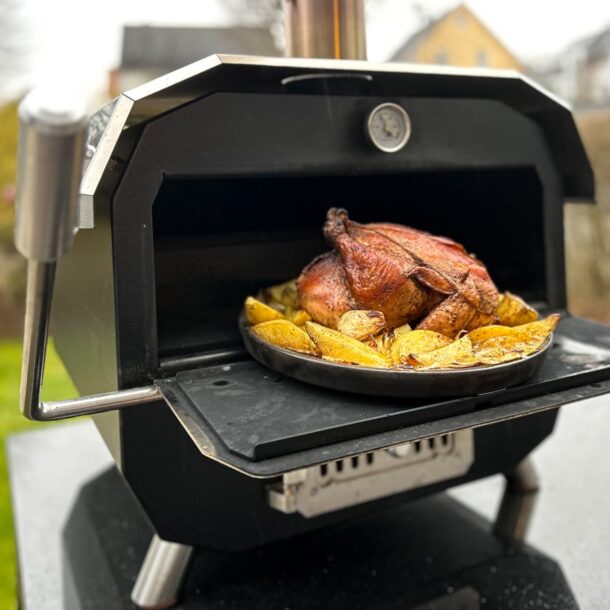 Ein kompakter, schwarzer Outdoor-Backofen mit geöffneter Klappe. Auf einer runden, dunklen Backplatte gart ein ganzes Brathähnchen, umringt von knusprigen Kartoffelspalten; im Hintergrund ein Garten/Hausszenerie Merklinger Bobby Keltenschale