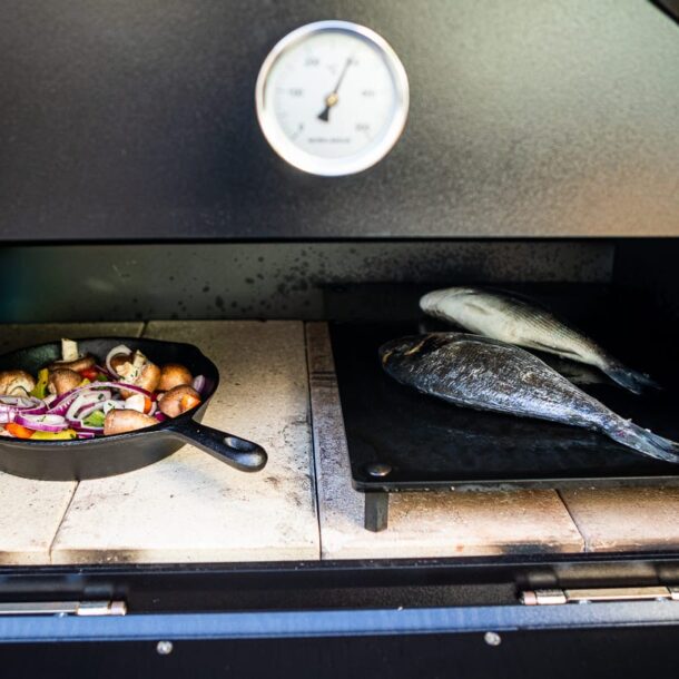 Zwei ganze Fische auf der Plancha, daneben Gusseisenpfanne mit Gemüse im Merklinger Ofen.