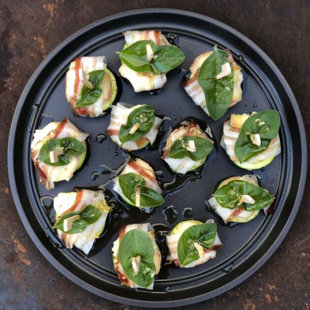 Zucchini-Häppchen: Runde Platte mit Zucchinischeiben im Speckmantel, belegt mit Salbeiblättern und Knoblauch, mit Olivenöl beträufelt