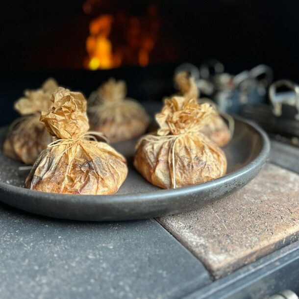 Im heißen Ofen liegende runde Platte mit kleinen, zu Päckchen gebundenen Backpapier-Beuteln; dahinter flackert die Flamme. Merklinger Zubehör Denkkeramik Keltenschale 03